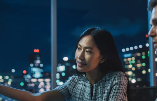 Financial Analyst Talking to Investment Banker in Late Evening at Work. Chatting About Real-Time Stock Chart Data on Tablet Computer. Businesspeople Have a Meeting in Broker Agency City Office.
