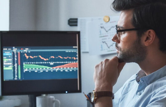 Busy working day. Side view of successful trader or businessman in formal wear and eyeglasses working with charts and market reports on computer screens in his modern office. Stock broker. Forex market. Trade concept