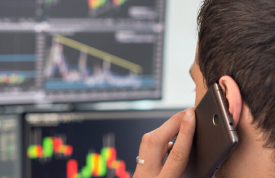 Over the shoulder view of and stock broker trading online while accepting orders by phone. Multiple computer screens ful of charts and data analyses in background.
