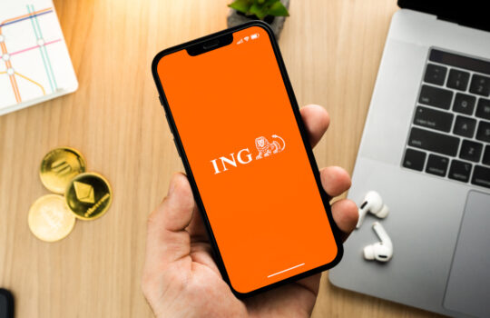 Man holding a smartphone with ING bank app on the screen on wooden table with a cryptocurrencies coins and computer. Office environment. Rio de Janeiro, RJ, Brazil. May 2022.