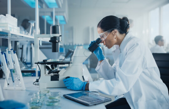 Medical Development Laboratory: Caucasian Female Scientist Looking Under Microscope, Analyzes Petri Dish Sample. Specialists Working on Medicine, Biotechnology Research in Advanced Pharma Lab