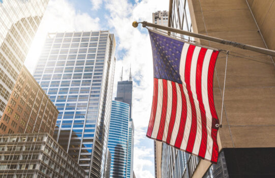USA flag in Chicago with with skyscrapers on background. American flag waving in the city on a sunny day. Clouds reflections on buildings facade