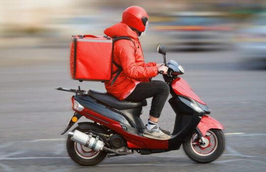 Food delivery moto scooter driver with red backpack behind back is on his way to deliver food. Courier on scooter delivering food. Quick shipping of goods to customers