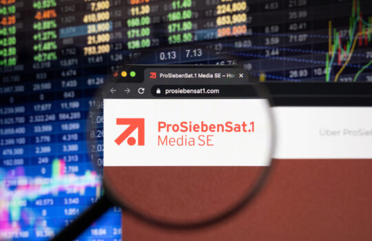 ProSiebenSat.1 Media company logo on a website with blurry stock market developments in the background, seen on a computer screen through a magnifying glass