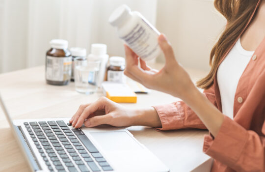 Wellness and dieting asian young woman, girl working from home using computer, typing or searching prescription on medicine label about vitamins information online, holding bottle of food supplement.