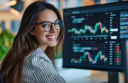 A smiling young businesswoman with glasses is sitting behind a desk looking at a monitor with a stock market graph monitoring market prices. widgets displaying the weather and the news daily schedule