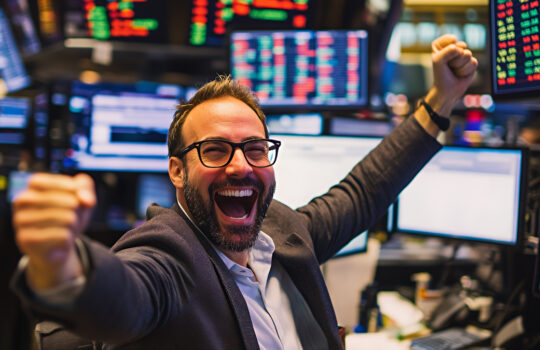 A successful businessman in a suit celebrating a win in front of stock market data screens with a joyful expression, fist in the air and a big smile.