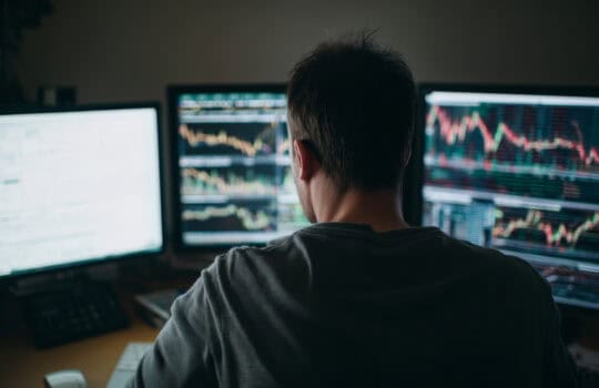 Behindtheshoulder view of male forex trader analyzing multiple currency charts on glowing monitors in dark home office.