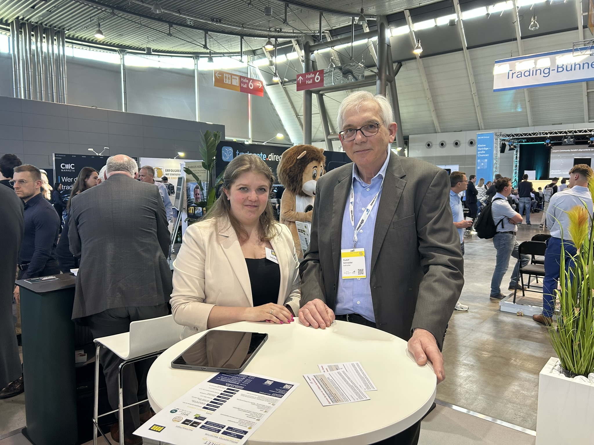 Two professionals at a trade fair booth, a woman in a beige blazer and a man in a gray suit standing by a round table with brochures and a tablet on it.