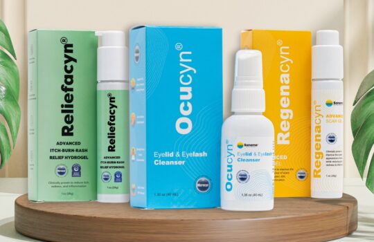 A row of eye-care and skin-relief products on a round wooden platform, including Ocucyn eyelid & eyelash cleanser, Reliefacyn hydrogel, and a yellow Regencyn box, with green plants on the sides.