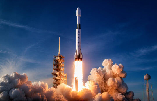 SpaceX rocket launching from a pad with fiery exhaust and billowing smoke against a blue sky.