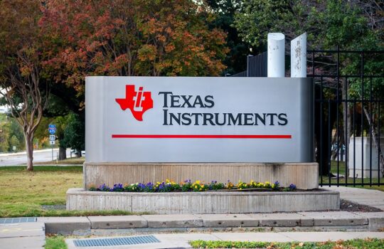 Texas Instruments sign on a concrete planter with flowers, behind a black fence and trees in autumn foliage.