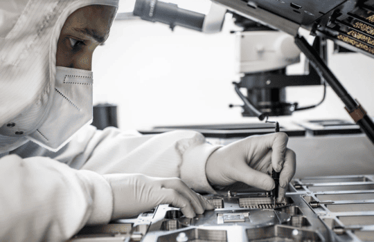 Lab technician in a cleanroom suit tools near an open circuit board, assembling microelectronics under a microscope