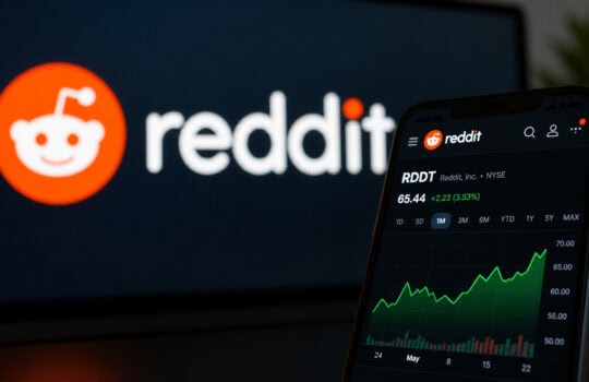 Close-up of a smartphone showing the Reddit app with a green upward-trending stock chart and price, blurred Reddit logo on a dark screen in the background, conveying investment focus.
