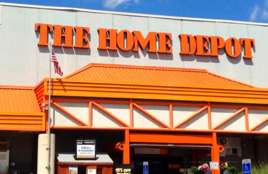 Exterior view of The Home Depot store with large orange sign and orange awning against a blue sky.