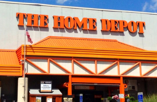 Storefront with large orange THE HOME DEPOT sign and orange architectural accents against a blue sky misted with clouds.