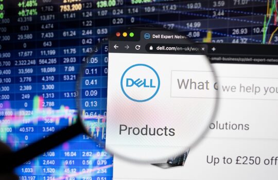 Close-up of a magnifying glass over a Dell webpage showing the Dell logo and 'Products' menu with a stock market screen in the background, suggesting a product search context.