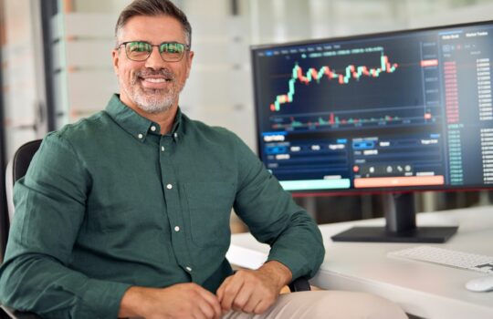 Smiling man in a green shirt seated at a desk with a monitor displaying stock charts and trading data, in a bright office.