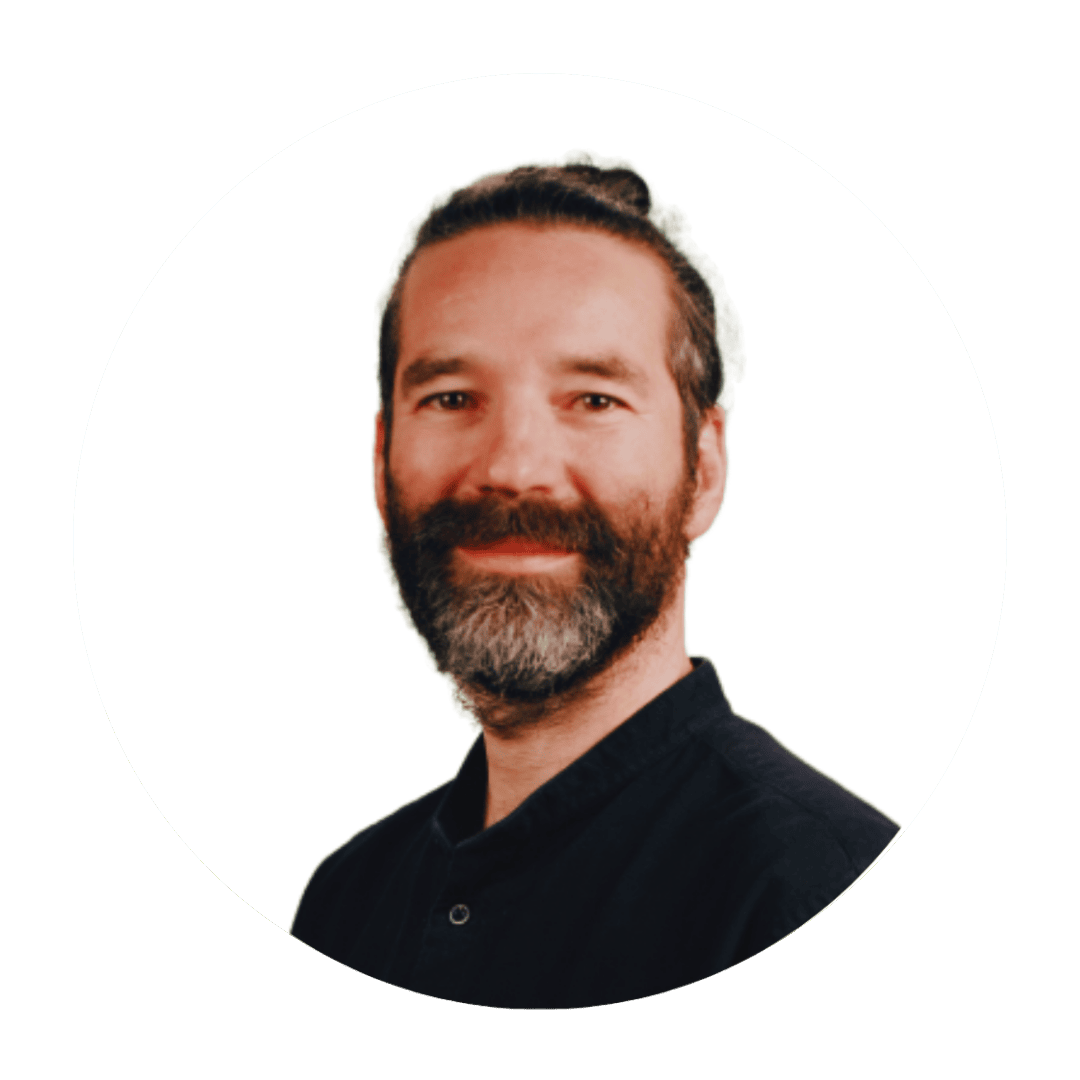 Portrait of a smiling man with a dark beard and bun, wearing a black shirt, against a white background.