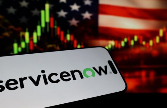 ServiceNow logo on a curved white board in the foreground with a blurred U.S. flag and stock chart in the background, suggesting business technology and finance.