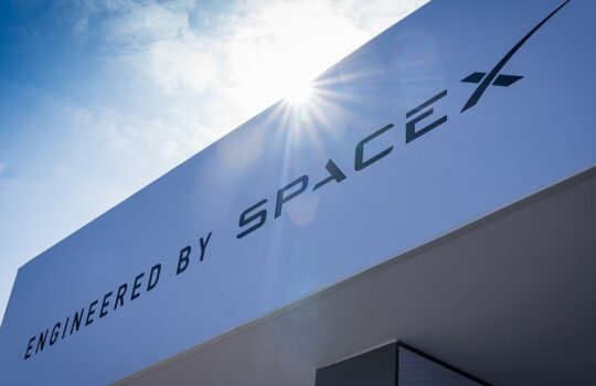 White exterior wall with a large 'Engineered by SpaceX' sign under a bright sun flare in a blue sky.