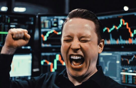 Trader celebrating with raised fist in front of multiple stock charts on monitors behind him.