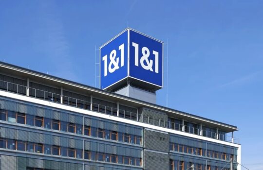 Modern office building with a large blue 1&1 sign on the rooftop against a clear blue sky.