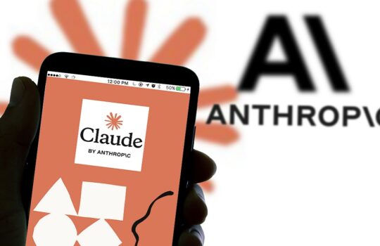 Smartphone held in hand showing the Claude by Anthropic app on a peach smartphone screen with geometric shapes in the foreground and an AI logo in the background.