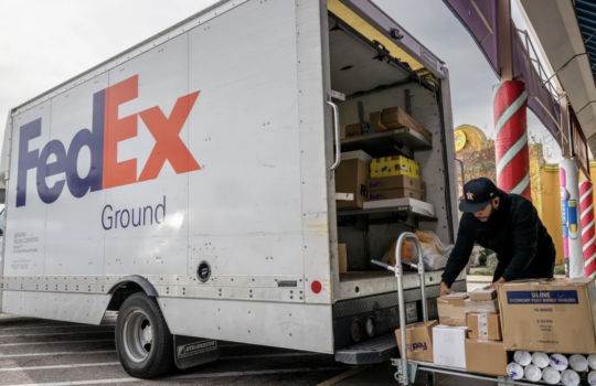 FedEx Ground delivery truck with open rear door, a worker stacking and loading boxes onto a dolly outside the truck