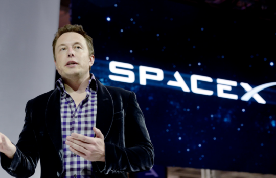 Man in a dark blazer and checkered shirt speaks on stage with a SpaceX backdrop.