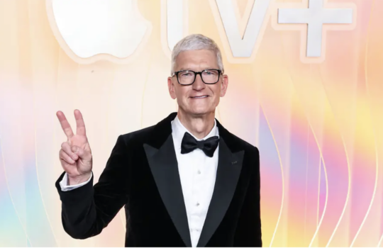 Man in a black tuxedo and glasses smiles at the camera, flashing a peace sign against a pastel, wavy backdrop.