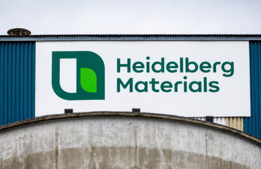 White sign on a blue industrial building reading 'Heidelberg Materials' with a green logo on the left side of the panel.