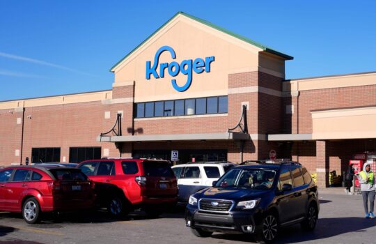Kroger supermarket storefront with a blue sign, brick facade, and parked cars in a sunny lot.
