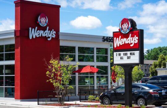 Exterior of a Wendy's fast-food restaurant with red branding, glass windows, and a parking lot on a sunny day.