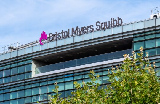 Bristol Myers Squibb corporate building with glass facade and trees in the foreground on a sunny day.