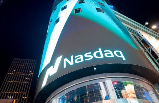 Night shot of a curved building displaying the illuminated Nasdaq logo at night, with blue lighting.