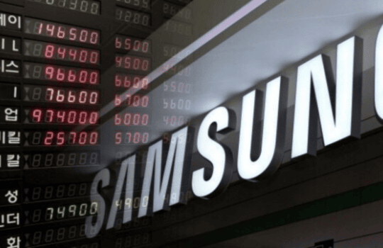 Samsung storefront sign with large white letters beside a scrolling stock ticker board displaying red and green numbers in Korean characters outside a financial building.