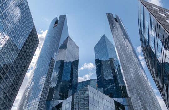Low-angle view of blue-tinted glass skyscrapers reflecting the sky, converging toward the center of a modern cityscape