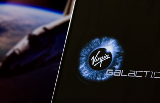 Close-up of a smartphone or device screen displaying the Virgin Galactic logo with a blue iris design and a space background nearby.