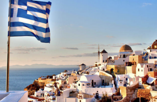 Greek flag waving above whitewashed Cycladic houses along a cliff with the sea in the background at sunset