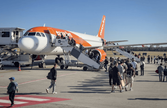 Passenger jet in orange livery being boarded with stairs and jet bridge at airport gate, crowds walking nearby