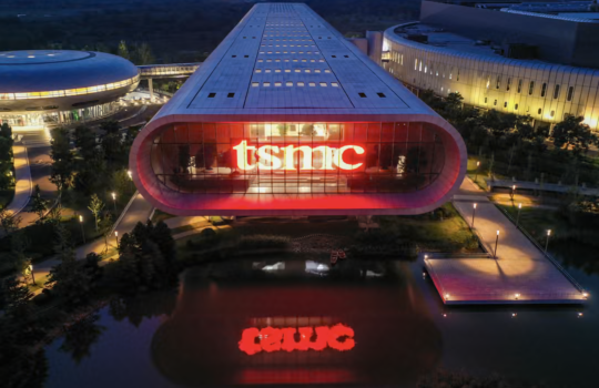 Night aerial view of a modern campus building with a curved glass front and a bright red 'tsmic' sign, reflected in the surrounding pond. The scene includes a circular building on the left and illuminated walkways.