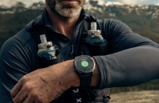 Close-up of a man's wrist wearing a rugged smartwatch showing a green connectivity icon, outdoors with a mountainous landscape in the background.