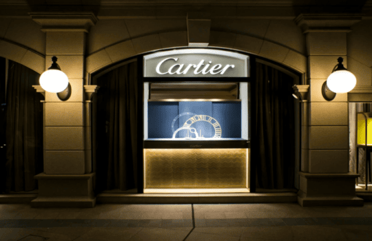 Cartier storefront at night with illuminated sign and a display window featuring a large clock gear design.