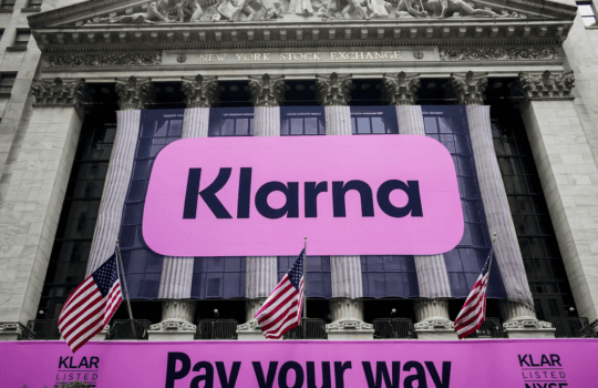 Front façade of the New York Stock Exchange with a large pink Klarna banner and American flags.
