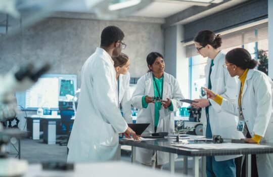 Group of five researchers in white lab coats collaborating around a bench with equipment in a modern laboratory, discussing a small device.