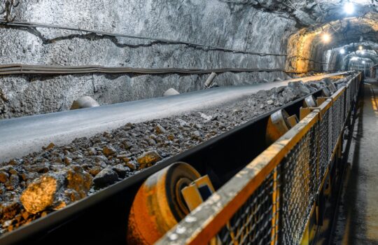 Underground mine tunnel with a running conveyor belt carrying rocky ore and a safety railing along the side.