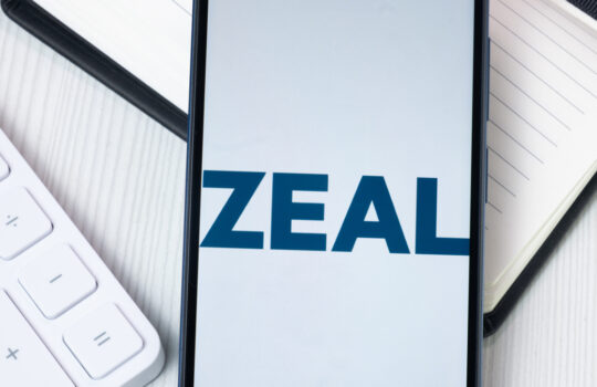 Smartphone screen shows the word ZEAL with a white keyboard and a lined notebook nearby on a light desk surface.
