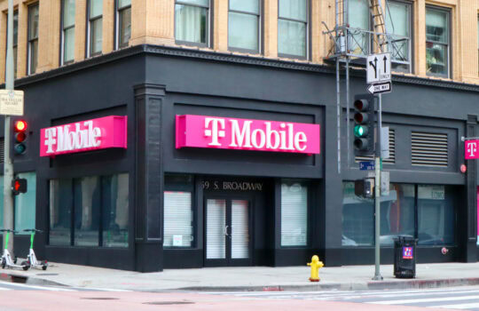 T-Mobile storefront on a dark corner building with bright pink signs and a crosswalk in the foreground.