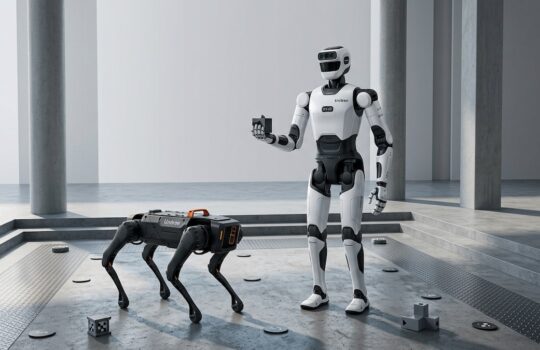 White humanoid robot stands in a gray testing chamber, holding a small card while a black four‑legged robot examines the floor nearby.